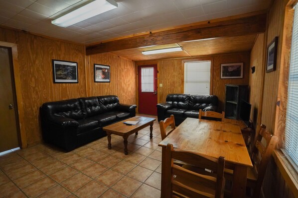 Une pièce aux murs en bois, avec deux canapés en cuir noir, une table à manger en bois et ses chaises, une table basse en bois et une porte rouge.