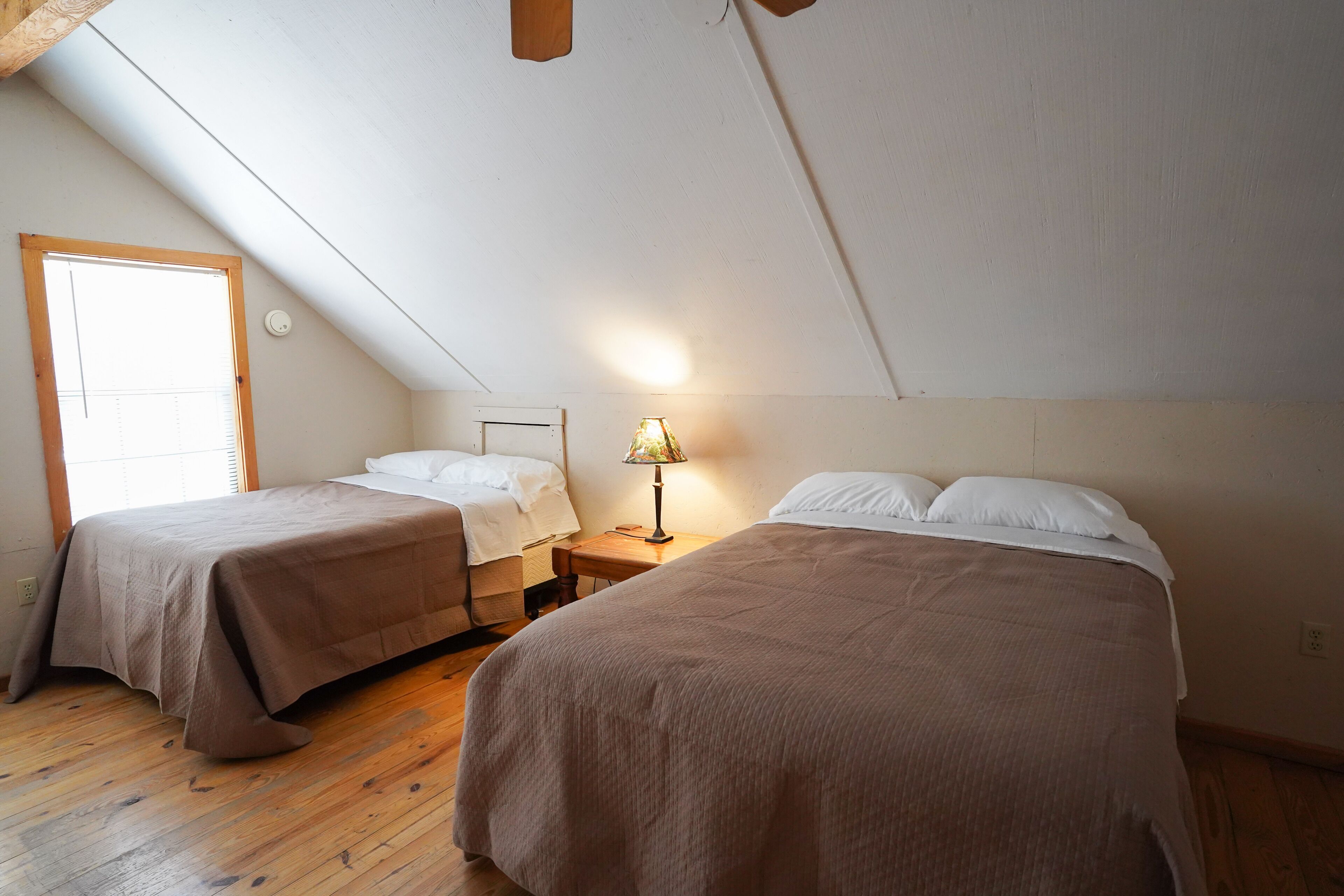 A bedroom with two beds, wooden flooring, a window, and a nightstand with a lamp.