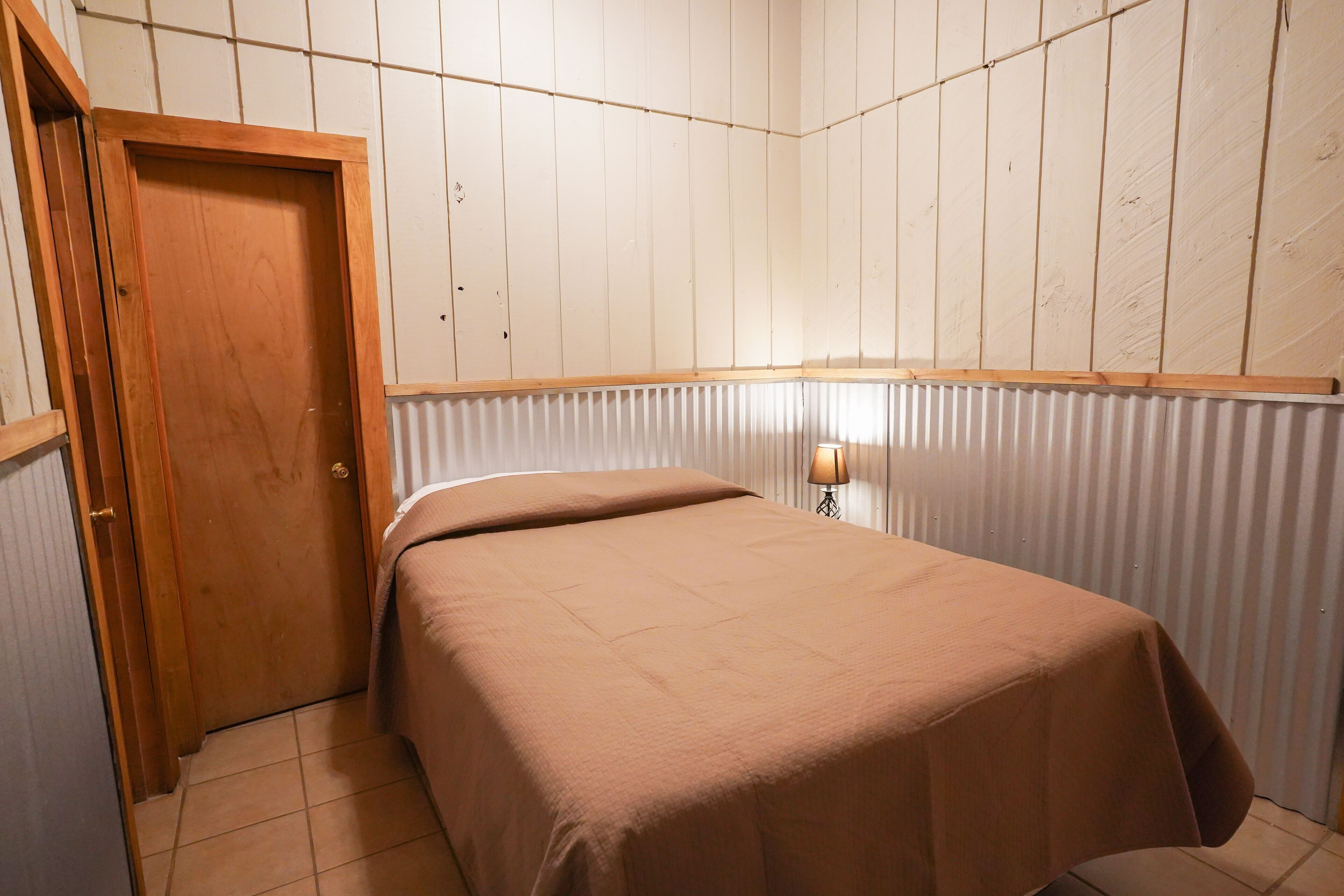 A bedroom with a wooden door, a bed with a brown bedspread, a wall-mounted lamp, and a radiator.