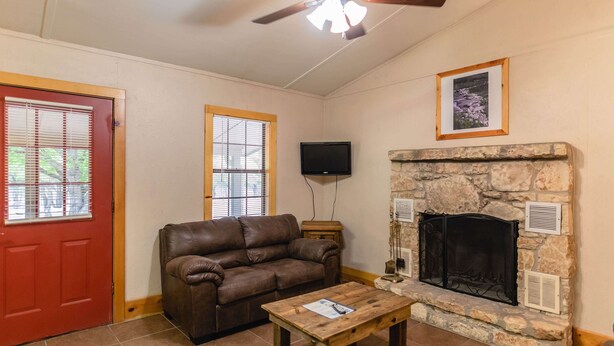 A living room with a stone fireplace, a brown leather sofa, a wooden coffee table, a ceiling fan, and a TV mounted on the wall. - Frio Country Resort - Within walking distance to the Frio River! (Concan)