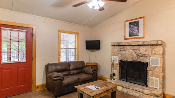 A living room with a stone fireplace, a brown leather sofa, a wooden coffee table, a ceiling fan, and a TV mounted on the wall. - Frio Country Resort - Within walking distance to the Frio River! (Concan)
