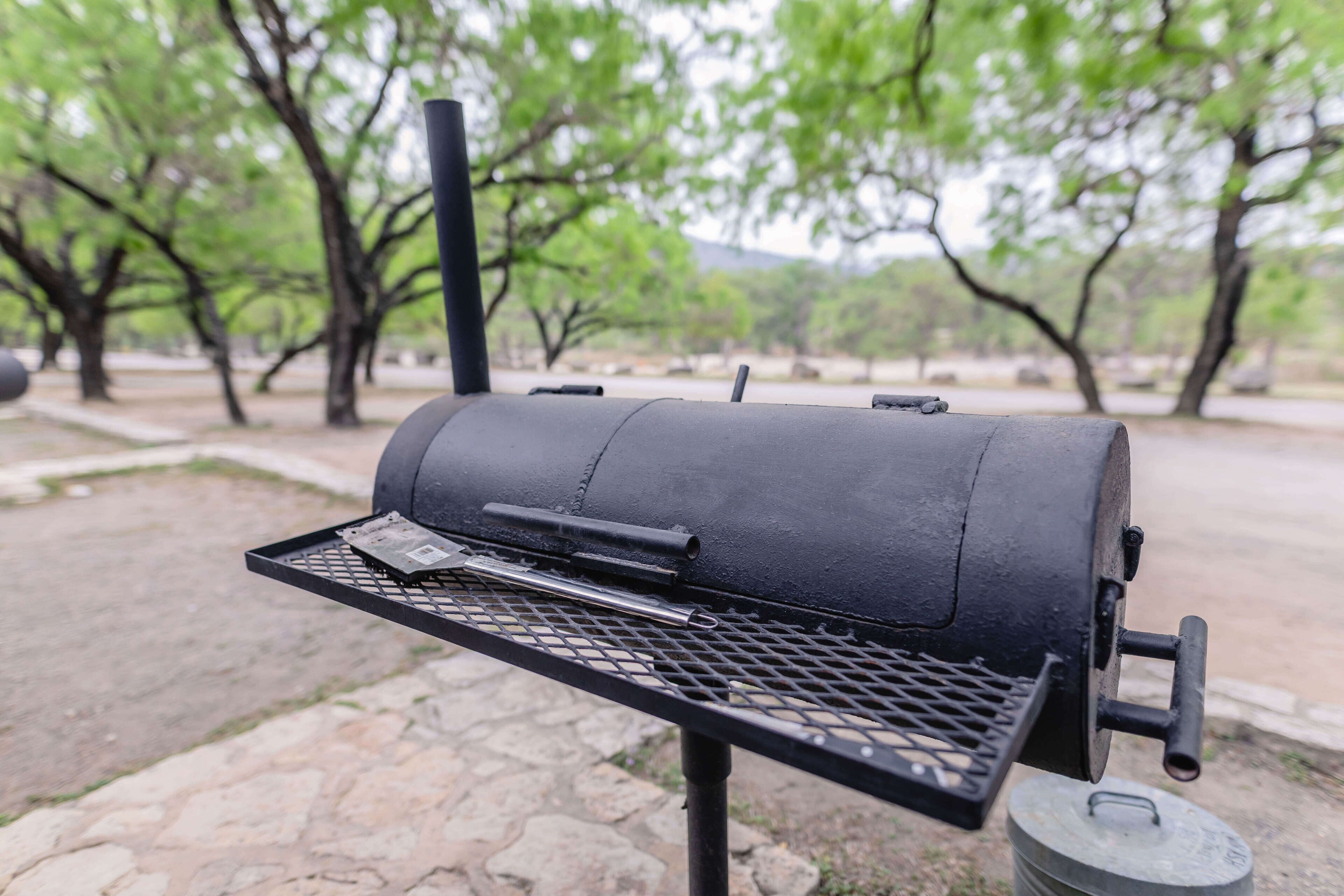 A black barbecue grill with a metal grate and a handle, set on a stone-paved area with trees in the background.
