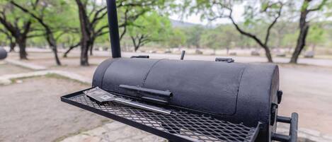 A black barbecue grill with a metal grate and a handle, set on a stone-paved area with trees in the background.