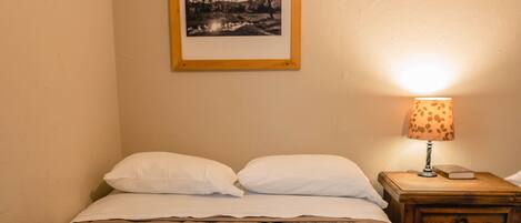 A neatly made bed with a white comforter and pillows, a wooden nightstand with a lamp and a book, and a framed picture on the wall.