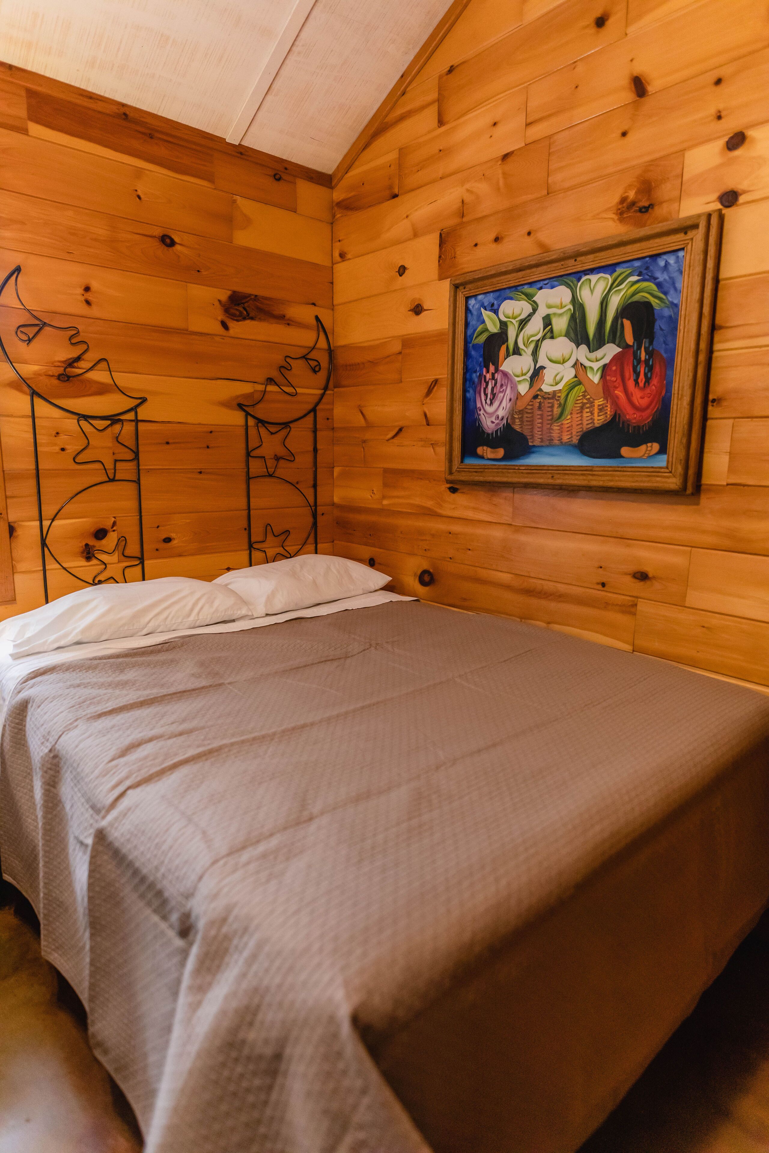A wooden cabin room with a bed, a painting on the wall, and a ceiling with a wooden beam.