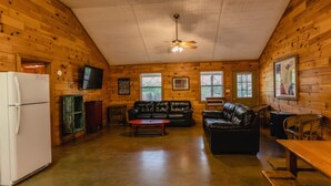 Interior de una cabaña de troncos con techo de madera, ventilador de techo, refrigerador, televisor, sofá de cuero, mesa de centro y mesa de comedor con sillas.