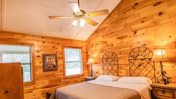 Un dormitorio tipo cabaña de madera con una cama, dos mesitas de noche, un ventilador de techo y una ventana con persianas.