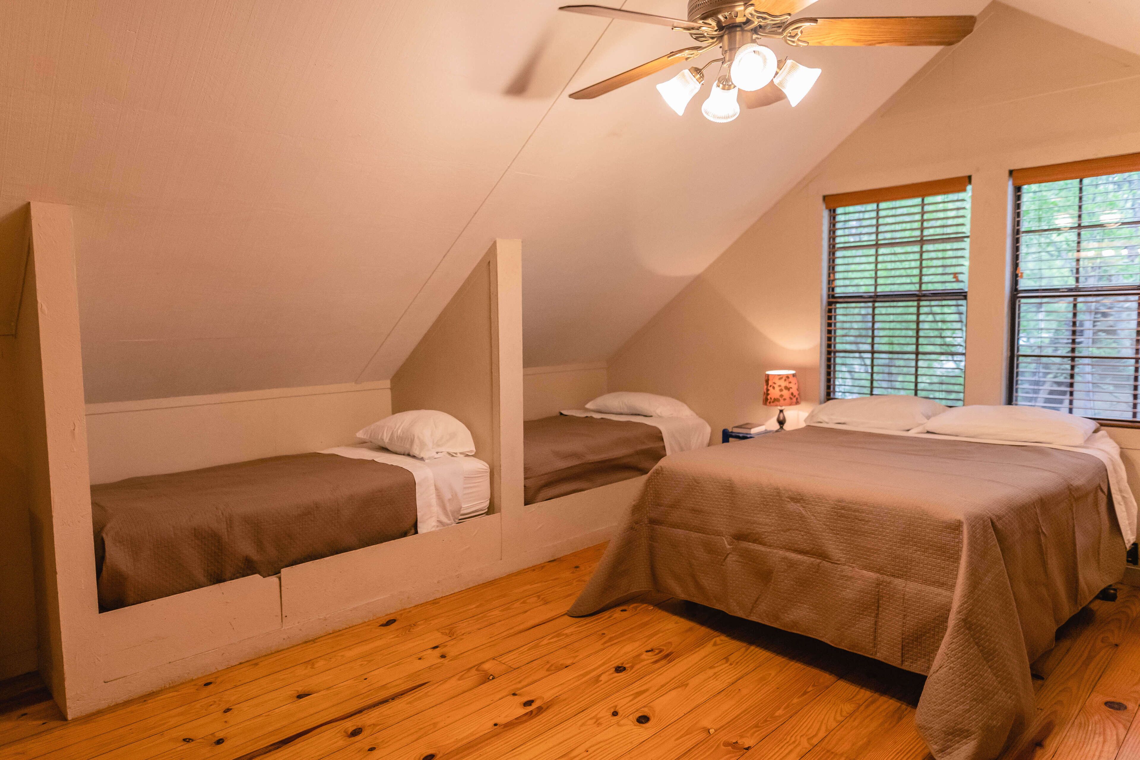 A room with two beds, wooden flooring, a ceiling fan, and windows with blinds.