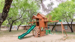 A playground with a slide and swings, surrounded by trees and buildings. - Frio Country Resort - Within walking distance to the Frio River! (Concan)
