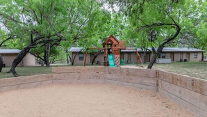 A playground with a wooden slide and swings, surrounded by sand and trees. - Frio Country Resort - Within walking distance to the Frio River! (Concan)