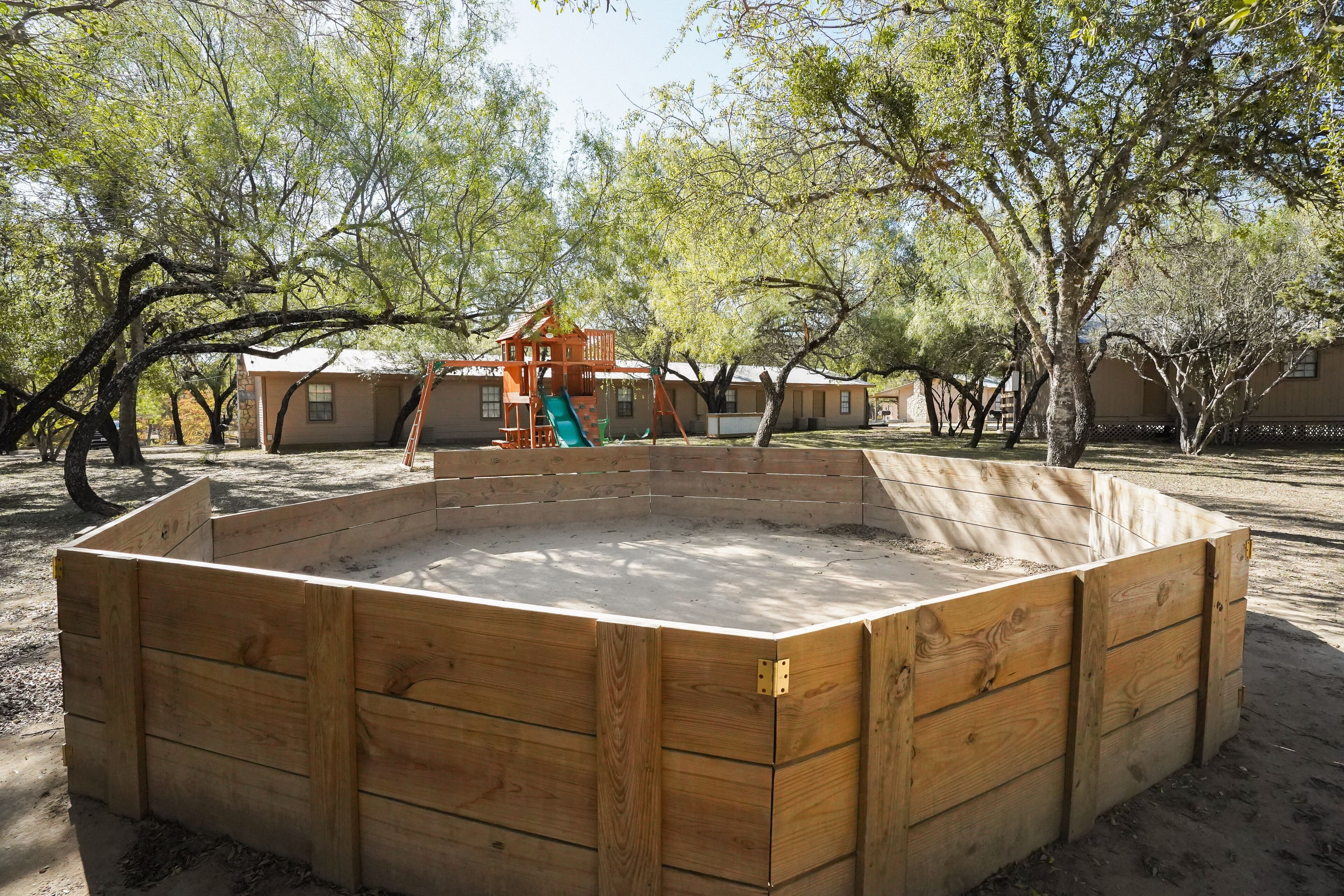 A wooden play structure with a slide and climbing area, surrounded by trees and a building in the background.