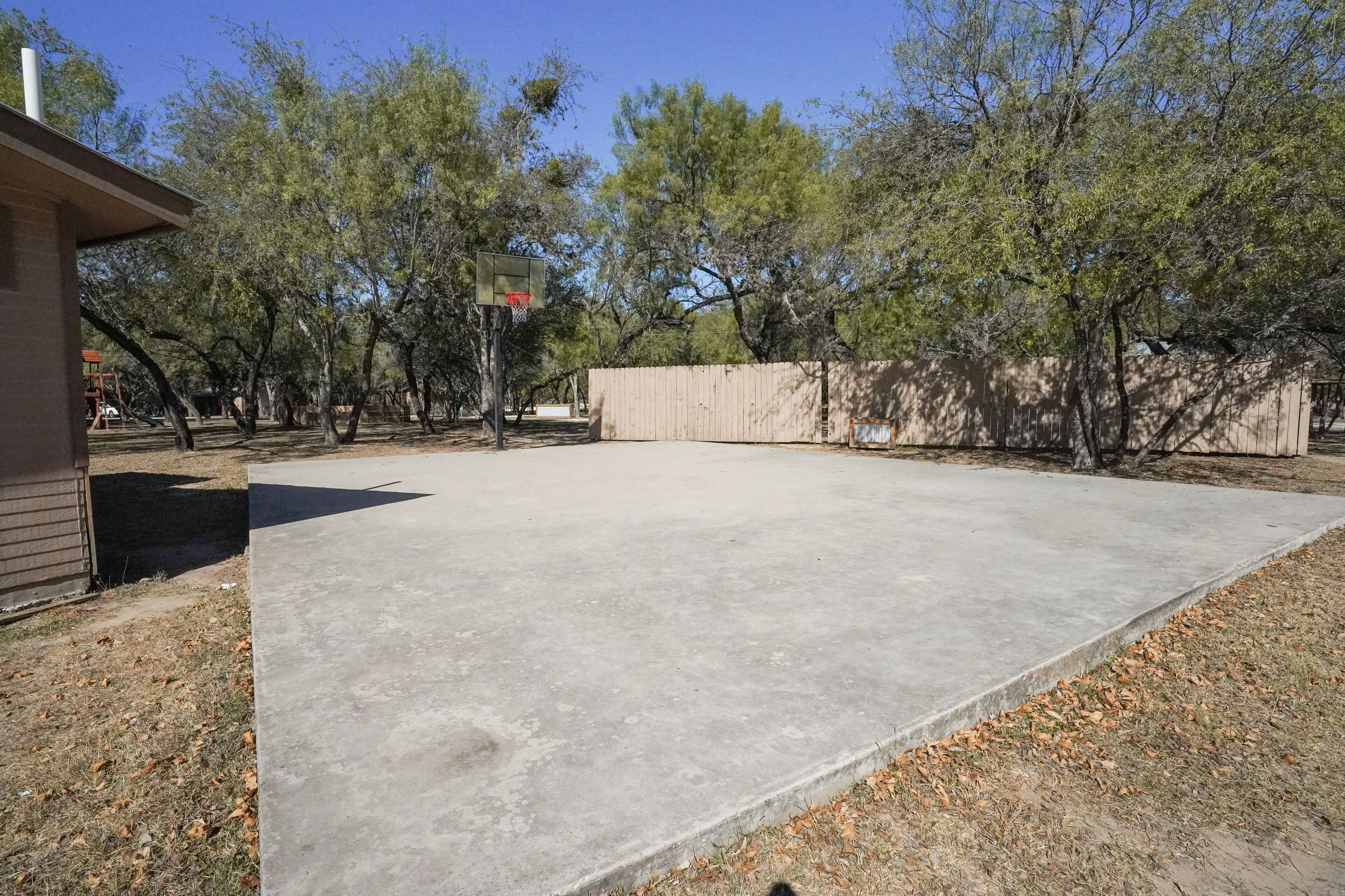 A concrete patio area with a basketball hoop, surrounded by trees and a wooden fence.