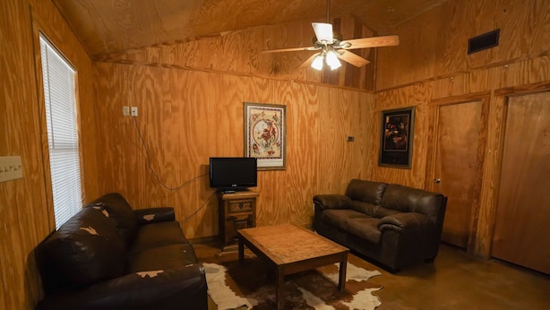A wooden-paneled room with a ceiling fan, a leather couch, a smaller sofa, a wooden coffee table, and a television on a stand. - Frio Country Resort - Within walking distance to the Frio River! (Concan)