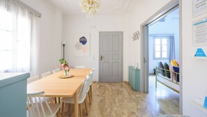 A dining area with a wooden table and white chairs, a chandelier, and a door leading to a bedroom with a bed and pillows.