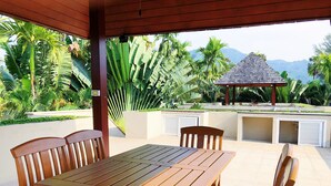 An outdoor patio with a wooden table and chairs, a covered seating area, and a view of lush greenery and mountains in the background. - Luxe 2br Pool Villa Walk to Bangtao Beach (Choeng Thale)