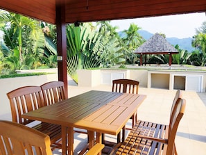 An outdoor patio with a wooden table and chairs, a covered seating area, and a view of a mountain and tropical vegetation.
