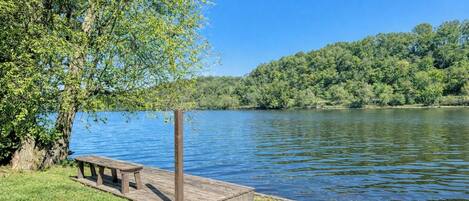 Une jetée en bois avec un banc, une zone herbeuse et un lac, avec des arbres en arrière-plan.