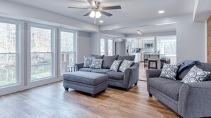 A spacious living room with grey sofas and ottomans, a ceiling fan, and large windows offering a view of the outdoors. - 4BR Lodge on Wolf Creek – Fishing, Hiking, New River Fun & Hokie Weekends (Narrows)