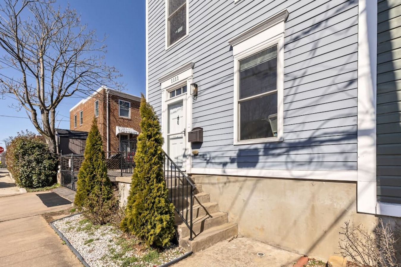 A two-story house with a front porch, a brick building adjacent to it, and a small garden with two evergreen trees.