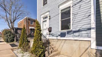 A two-story house with a front porch, a brick building adjacent to it, and a small garden with two evergreen trees.
