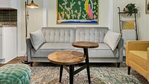 A living room with a grey sofa, two wooden side tables, a green ottoman, a patterned rug, a painting of a bird, a floor lamp, and a plant on a shelf. - 2bd1b Apart DT Safety Harbor, FREE Wifi (Safety Harbor)