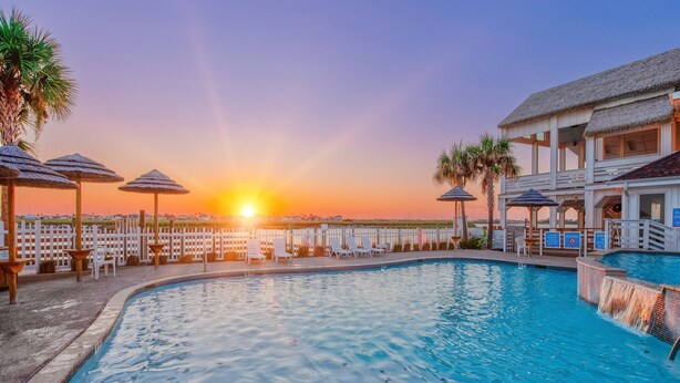 Ein Swimmingpool mit Liegestühlen und Sonnenschirmen, ein Gebäude mit einem Reetdach und ein Sonnenuntergang im Hintergrund.