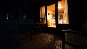 Une terrasse extérieure à la lumière tamisée, avec une table et des chaises, un banc, et une vue sur un bâtiment dont l’intérieur est éclairé à travers une fenêtre.