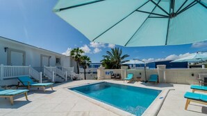A swimming pool with lounge chairs and umbrellas, surrounded by a white building with a staircase and palm trees.