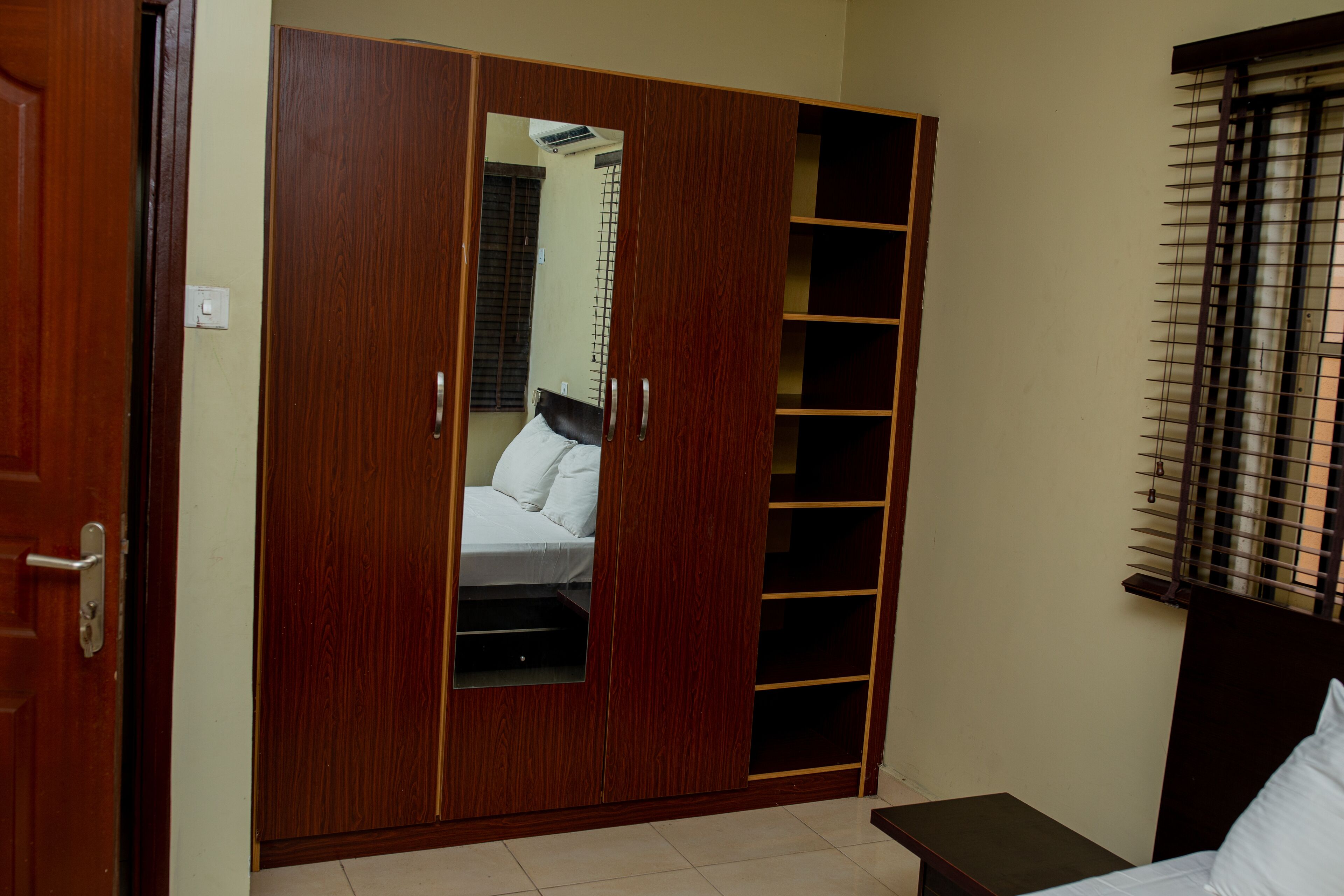 A bedroom with a wooden wardrobe, a bed, and a window with blinds.