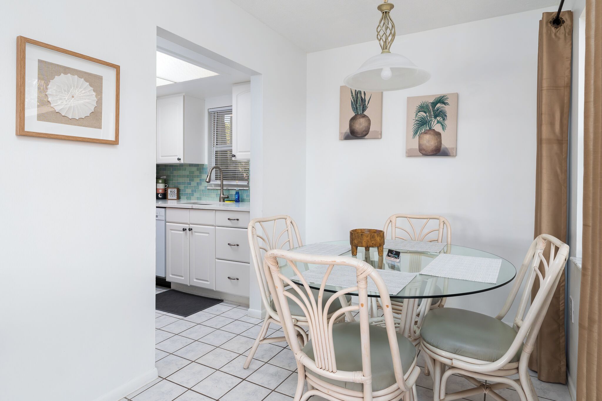 A dining area with a glass table and wicker chairs, a kitchen in the background, and two potted plants on the wall.