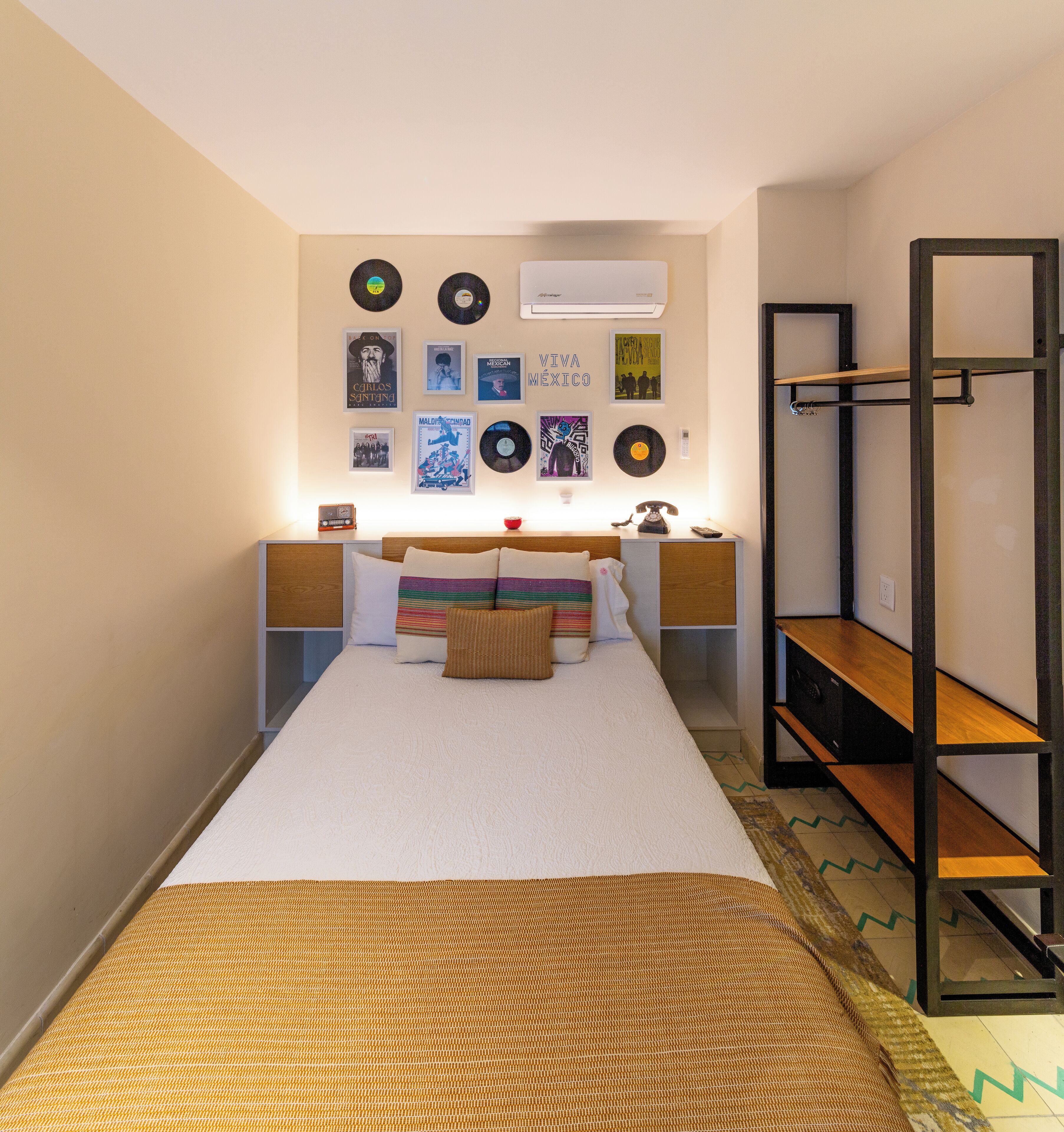 A bedroom with a bed, a bedside table, a wall-mounted shelf, and a wall with framed pictures and records.