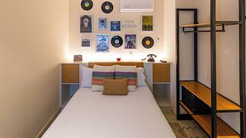 A bedroom with a bed, a bedside table, a wall-mounted shelf, and a wall with framed pictures and records.