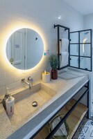 A modern bathroom with a large mirror, white tiled walls, a granite countertop, a towel rack, and a shower area.