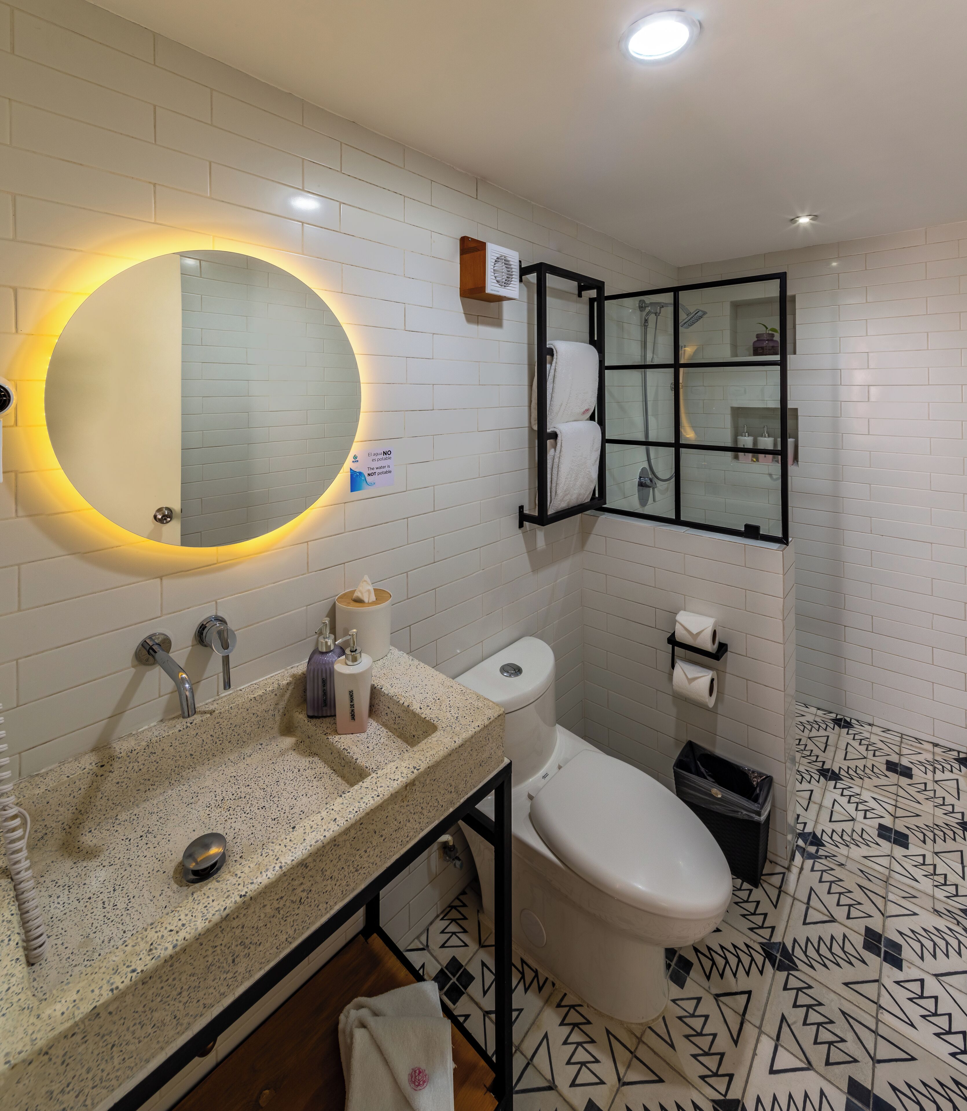 Un baño moderno con pared de azulejos blancos, un espejo redondo con luz amarilla, un inodoro, un lavabo con dispensador de jabón y un perchero para toallas.