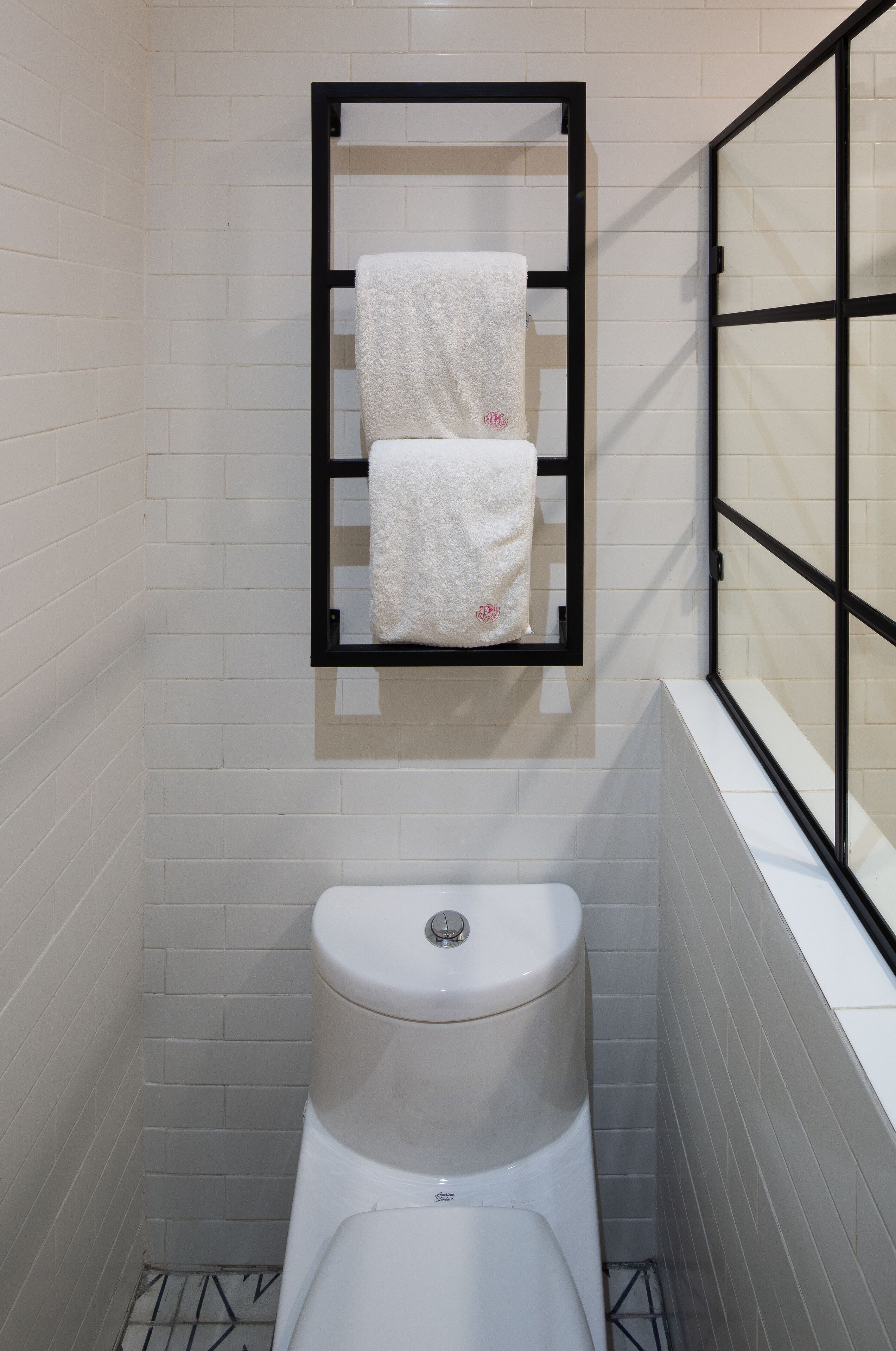 A small, modern bathroom with white tiled walls, a white toilet, and a black-framed mirror with two white towels hanging.