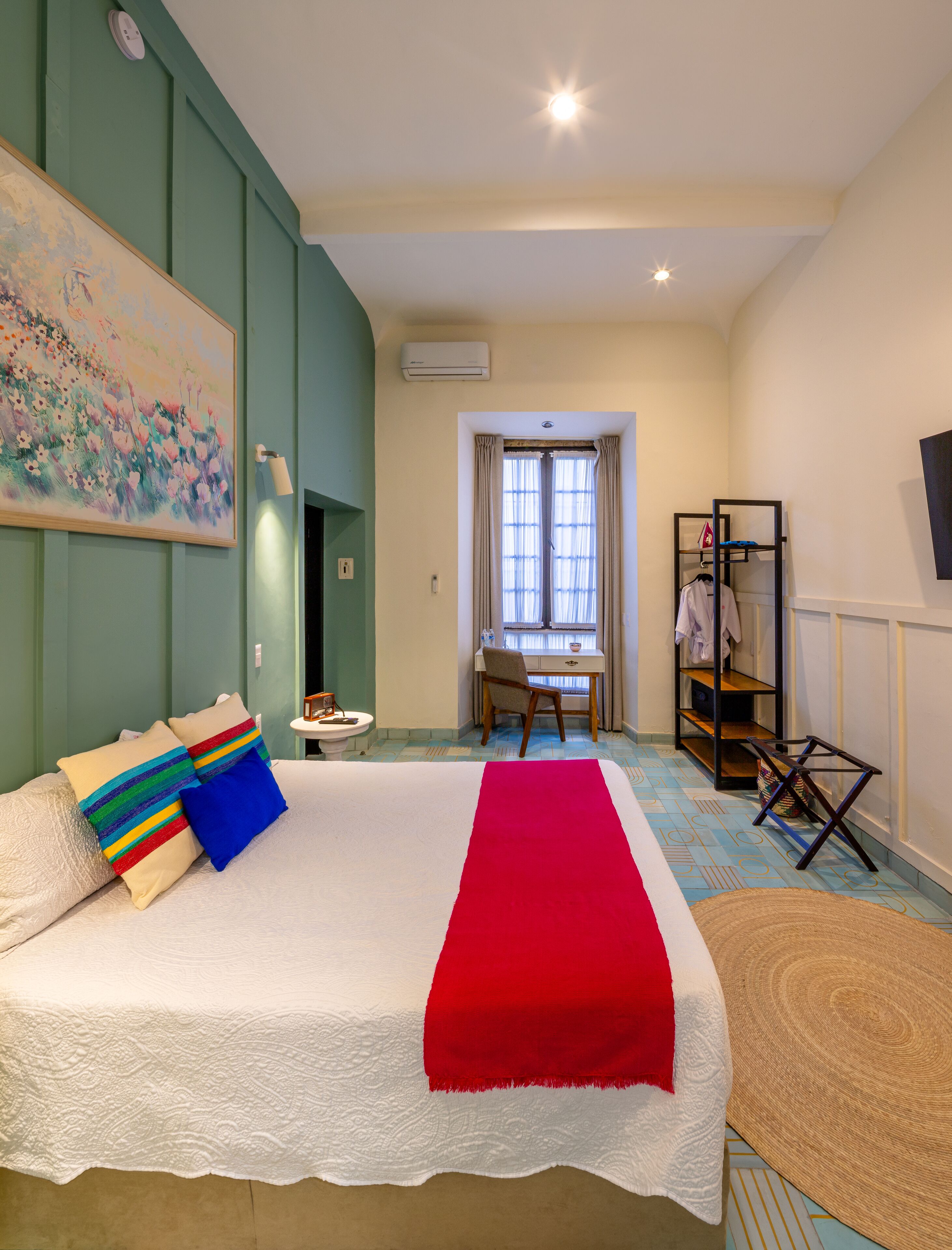 A bedroom with a large bed, a desk, and a chair. There is a painting on the wall and a window with curtains.