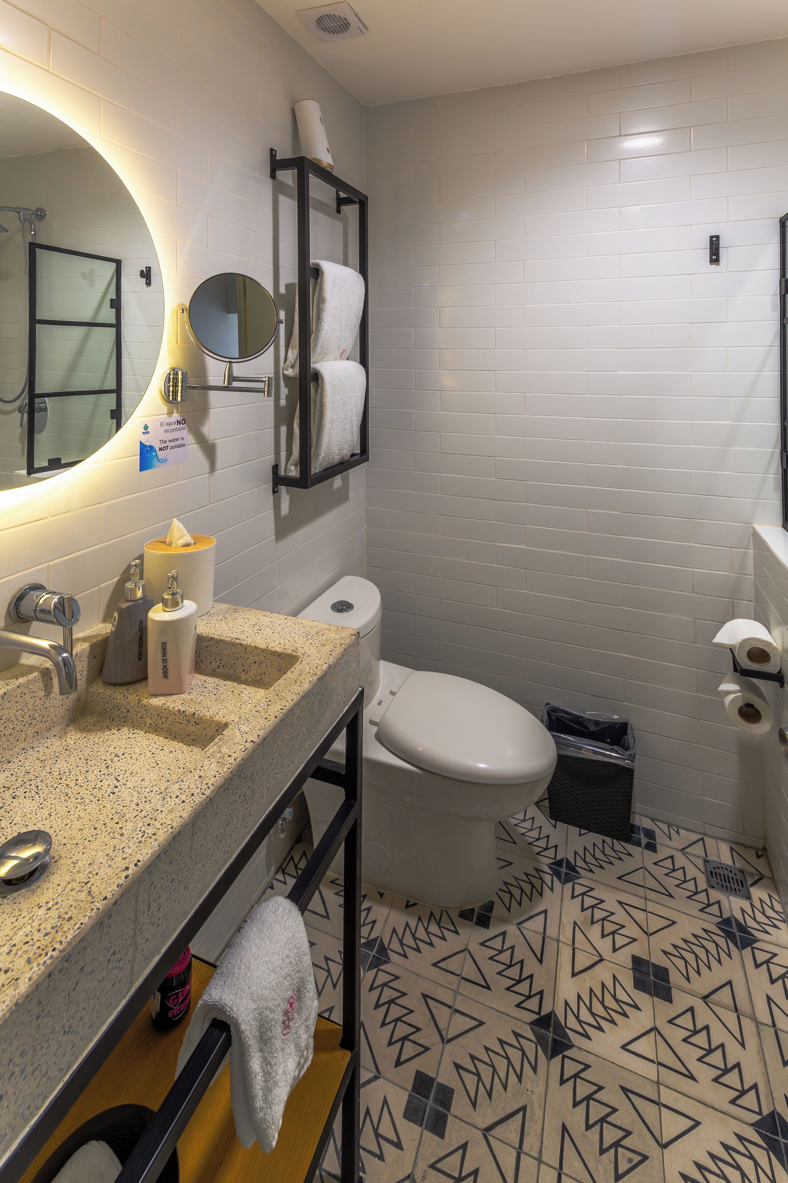 Un baño moderno con pared de azulejos blancos, un lavabo de granito, un espejo redondo, un perchero para toallas y un inodoro.