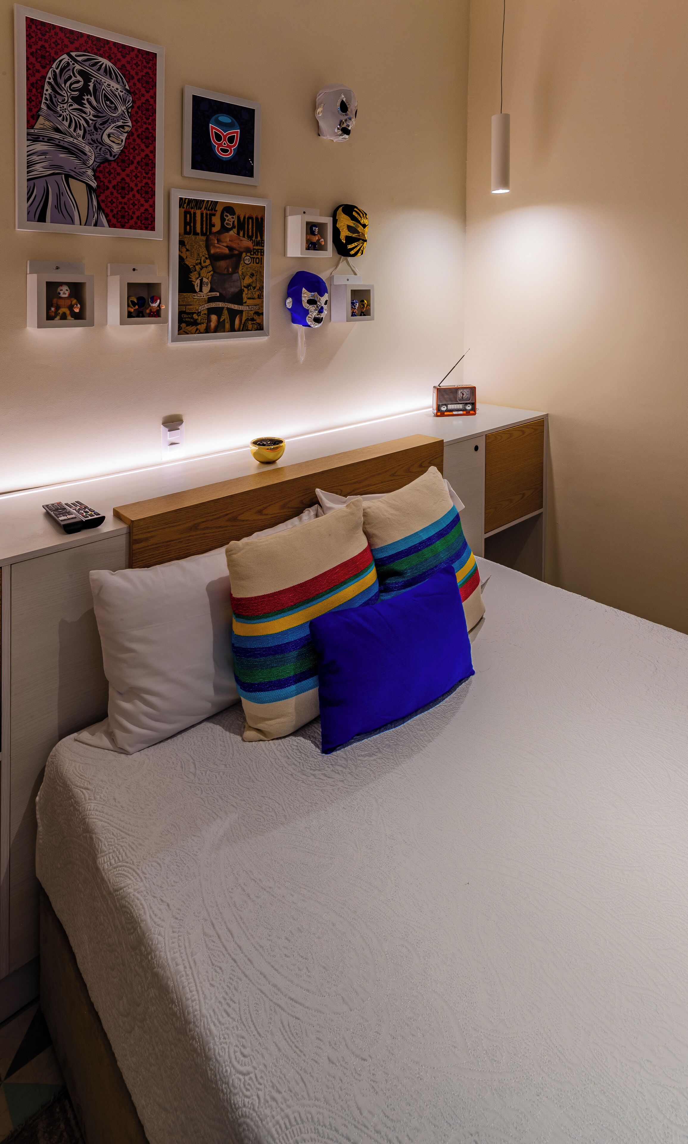 Una cama bien hecha con una colcha blanca y almohadas a rayas de colores, una mesita de noche con un radio y una pared decorada con marcos con fotografías y máscaras.