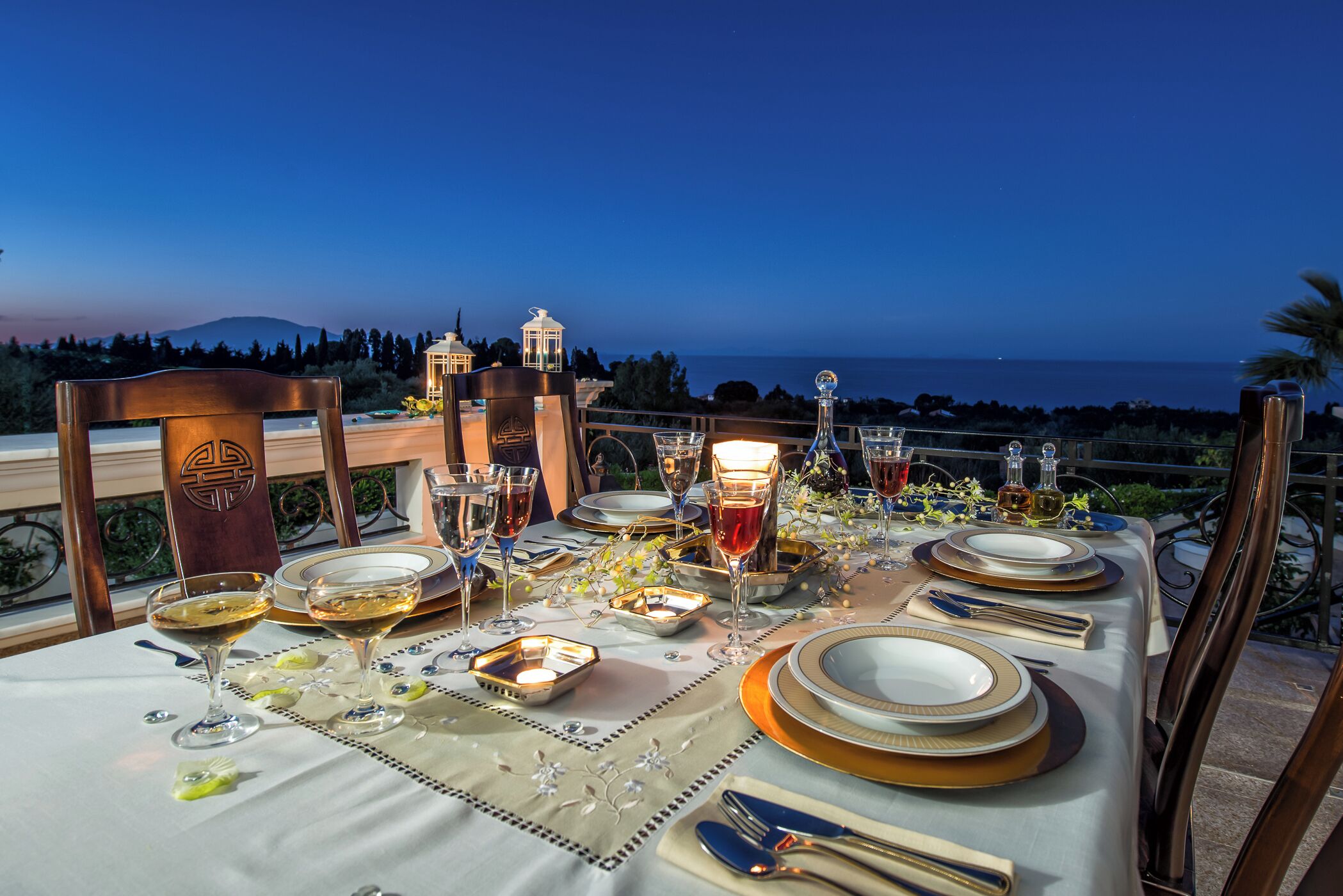 A table set for a meal with a view of the sea and mountains at dusk.