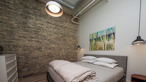 A bedroom with a brick wall, a bed with pillows, a bedside table, and a painting on the wall. - Flat 203 Front and Union (Traverse City)