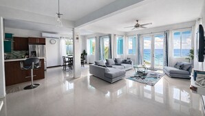 Uma sala de estar ampla com cozinha, espaço para refeições e grandes janelas com vista para o oceano.