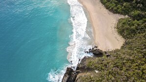 Vista aérea de uma área costeira com uma praia arenosa curvada, vegetação densa e falésias rochosas.