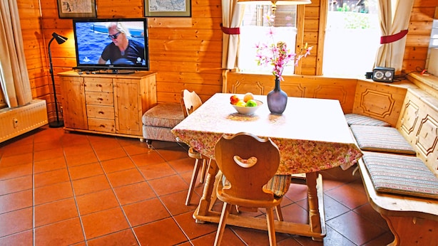 Interior de una cabaña de madera con una mesa de comedor, sillas, un televisor y una ventana con cortinas.