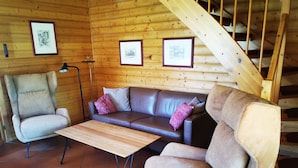 Interior de una cabaña de madera con un sofá, dos sillones, una mesa de centro de madera y una escalera.