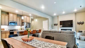 A modern kitchen with wooden cabinets, a dining area with a wooden table and chairs, a gray sofa, and a flat-screen TV mounted on the wall.