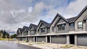 A row of modern, two-story houses with dark gray exteriors and pitched roofs. Each house has a garage and a front yard with small plants.