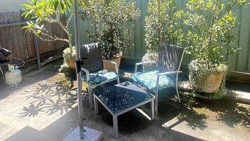 Een terras buiten met een tafel en stoelen onder een grote parasol, omringd door potplanten en een houten hek.