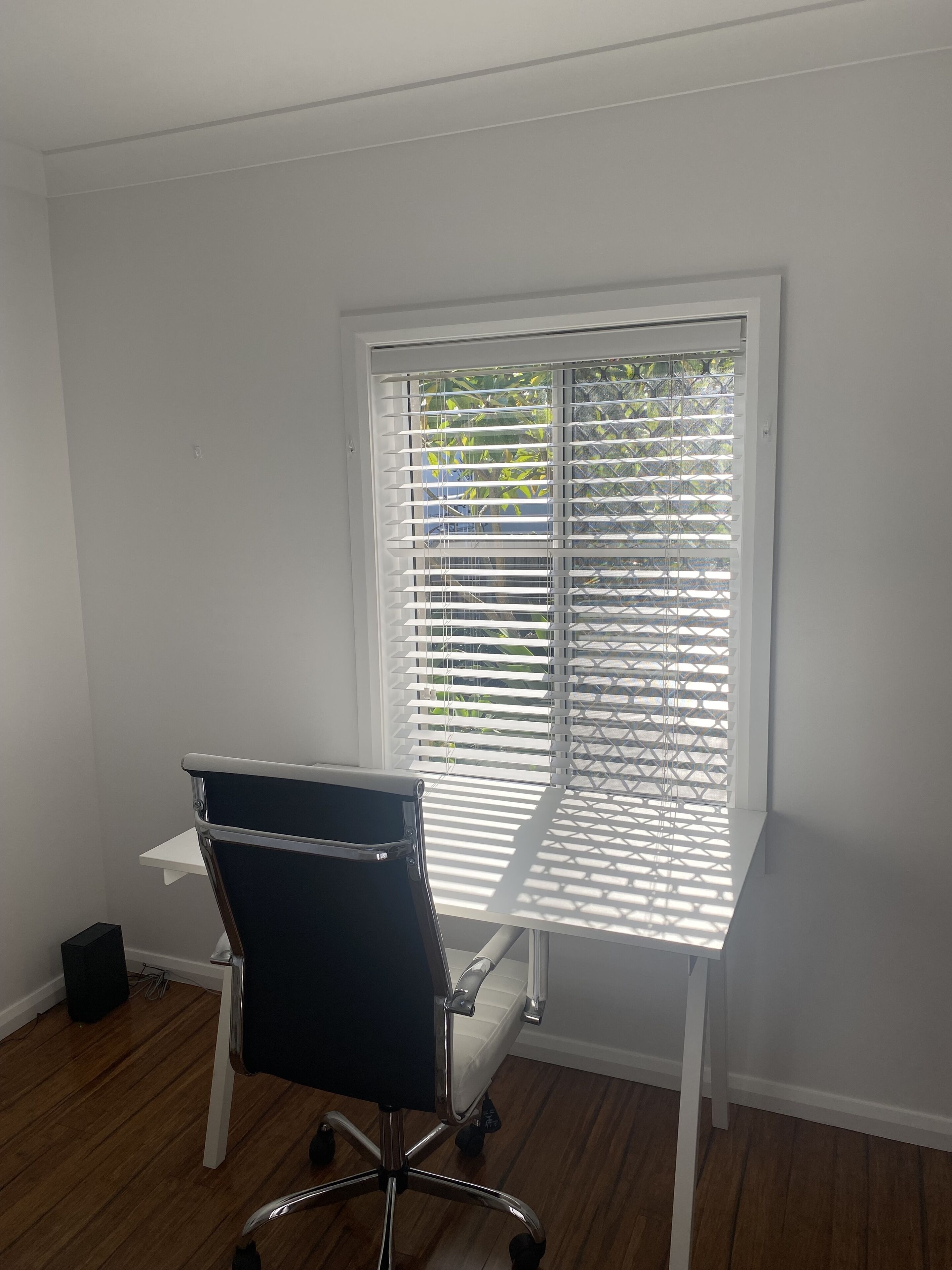 A room with a white desk, black office chair, and a window with white blinds.