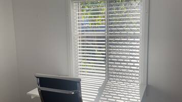 A room with a white desk, black office chair, and a window with white blinds.