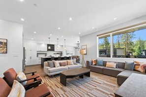 Ein modernes Wohnzimmer mit einer Couch, einem Couchtisch und Sesseln. Es gibt eine Küchenzeile mit Schränken und einem Esstisch. Das Zimmer verfügt über große Fenster mit Blick auf Bäume und Gebäude.
